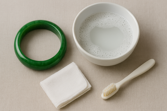 A green jade bangle rests on a beige fabric beside a bowl of soapy water, a white microfiber cloth, and a soft toothbrush—showing the essentials for safely cleaning and caring for jade jewelry.