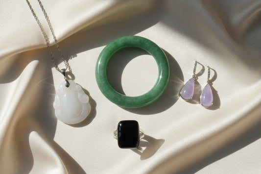 Collection of natural jade jewelry featuring green jadeite bangle, white nephrite pendant, lavender earrings, and black jade ring on silk background