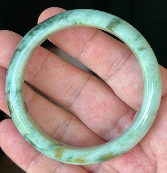 Hand holding a 55.3mm certified Grade A natural Burmese jadeite bangle in icy apple and forest green with black and honey hues, item JB543
