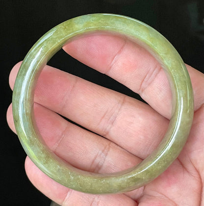 Hand holding a 58mm certified Grade A Burmese jadeite bangle in icy apple and forest green with milky honey white hues, item JB040