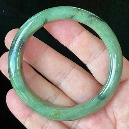 Hand holding a 58mm certified Grade A Burmese jadeite bangle in icy apple green with black floral design and off-white specks, item JB905