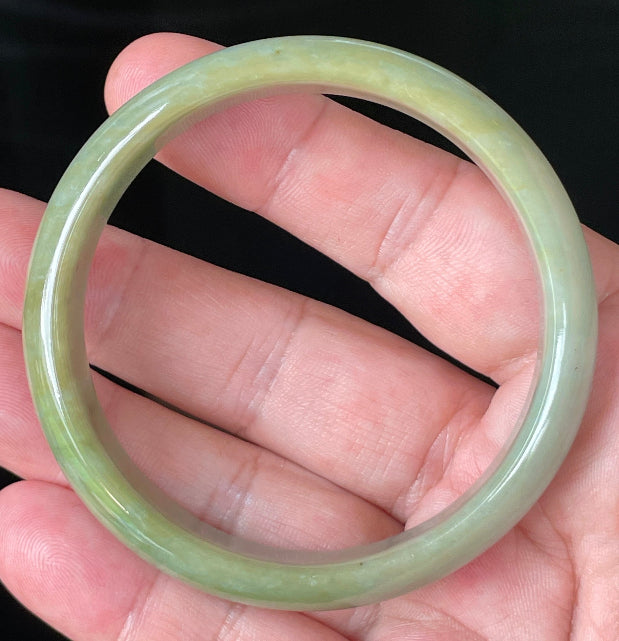 Hand holding a 59mm certified Grade A Burmese jadeite bangle in icy apple green and bluish milky white with honey hues, item JB213