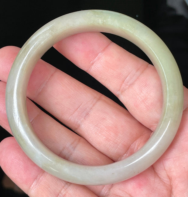 Hand holding a 58.5mm certified Grade A natural Burmese jadeite bangle in icy apple green, brown and greyish milky white hues, item JB091