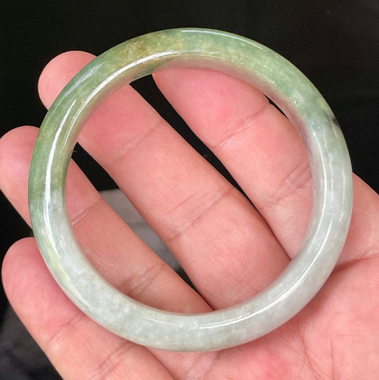 Hand holding a 54.8mm certified Grade A Burmese jadeite bangle in icy apple green with greyish milky white and black hues, item JB050