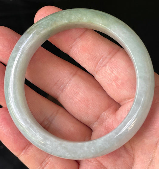 Hand holding a 56.2mm certified Grade A Burmese jadeite bangle in icy apple green and greyish milky white with light lavender hues, item JB203