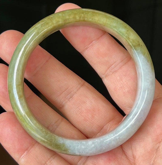 Hand holding a 58.6mm certified Grade A natural Burmese jadeite bangle in icy apple green and honey with greyish milky white tones, item JB521