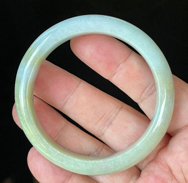 Hand holding a 57mm certified Grade A Burmese jadeite bangle in icy apple green with light lavender and milky white hues, item JB912