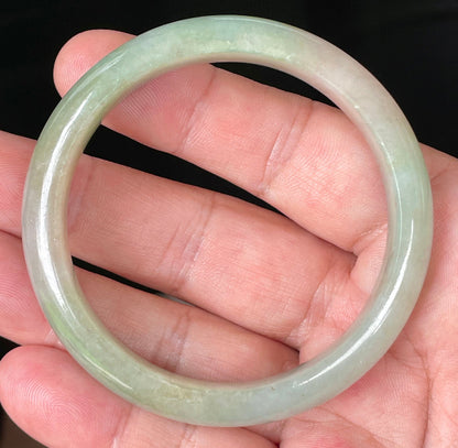 Hand holding a 57mm certified Grade A natural Burmese jadeite bangle in icy apple green and milky white hues, item JB079