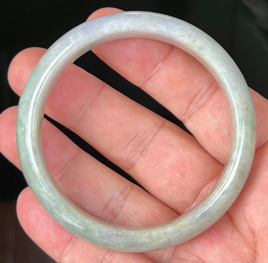 Hand holding a 57.8mm certified Grade A natural Burmese jadeite bangle in icy apple green with milky white and greyish hues, item JB532