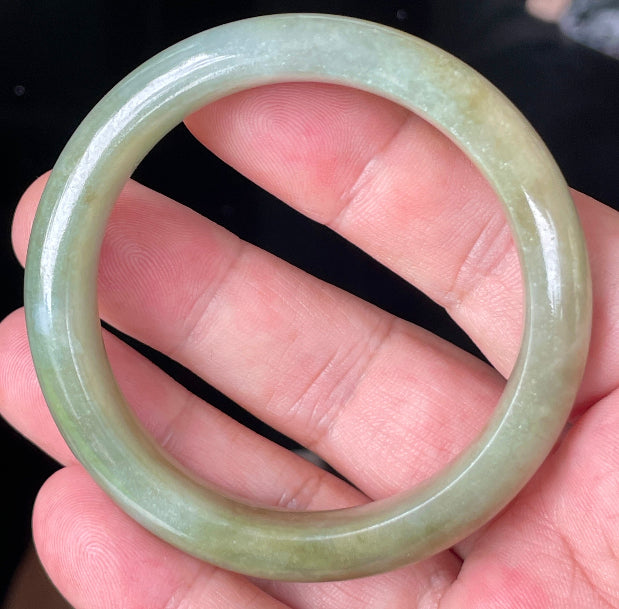 Hand holding a 53mm certified Grade A natural Burmese jadeite bangle in icy apple green with milky white and honey brown hues, item JB355