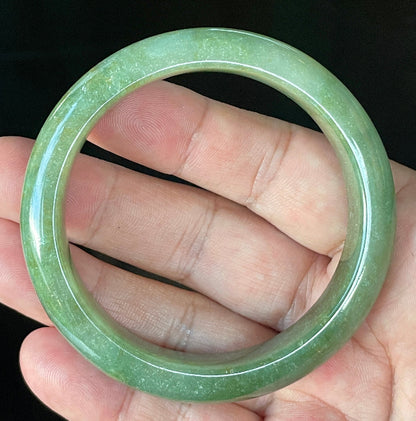 Hand holding a 57.6mm certified Grade A Burmese jadeite bangle in icy apple green with milky white and honey hues, item JB916