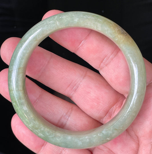 Hand holding a 58.5mm certified Grade A Burmese jadeite bangle in icy apple green with milky white and honey hues, item JB212