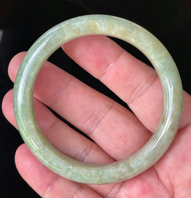 Hand holding a 58.2mm certified Grade A Burmese jadeite bangle in icy apple green, milky white, and honey brown hues, item JB830