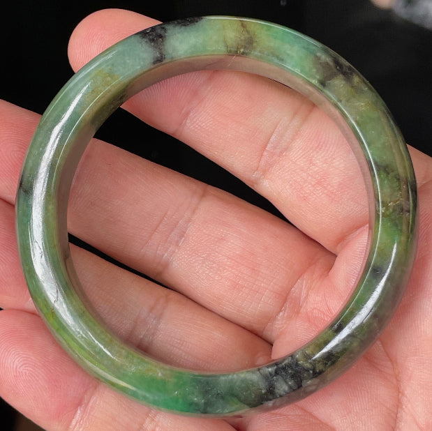 Hand holding a 54.5mm certified Grade A natural Burmese jadeite bangle in icy apple and imperial green with black and honey hues, item JB351