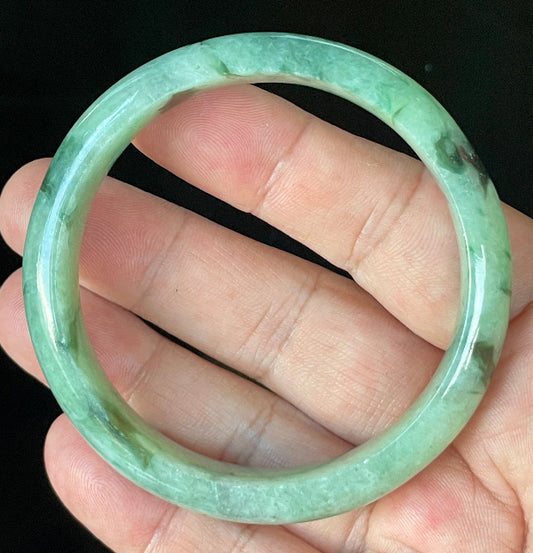 Hand holding a 57.5mm certified Grade A Burmese jadeite bangle in icy apple and imperial green with black and greyish milky white hues, item JB911