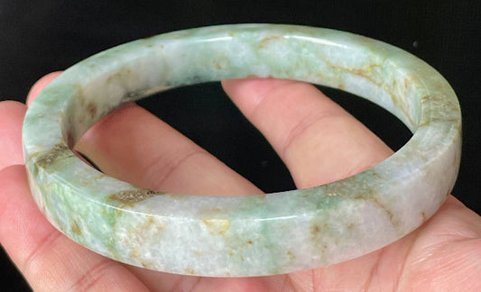 Hand holding a 77.9mm certified Grade A Burmese jadeite flat style bangle in icy apple, imperial, and moss green with milky white hues, item JB934