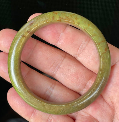 Hand holding a 55mm certified Grade A Burmese jadeite bangle in icy apple and moss green with honey red hues, item JB873