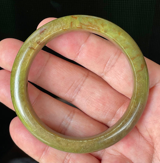 Hand holding a 55mm certified Grade A Burmese jadeite bangle in icy apple and moss green with honey red hues, item JB873