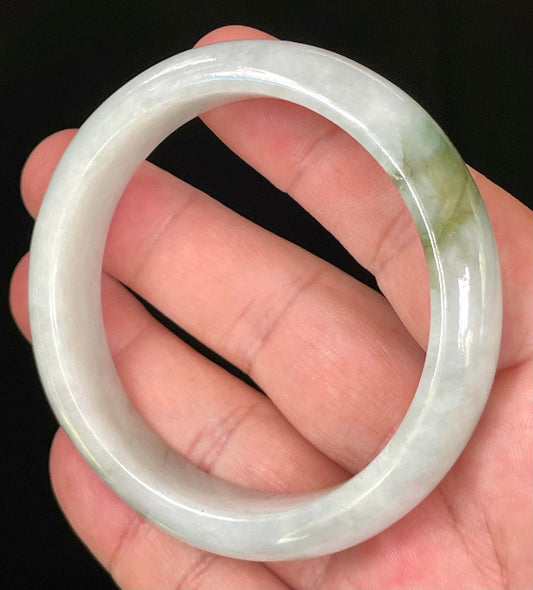 Hand holding a 57.5mm certified Grade A Burmese jadeite bangle in icy apple and moss green with light greyish milky white hues, item JB025