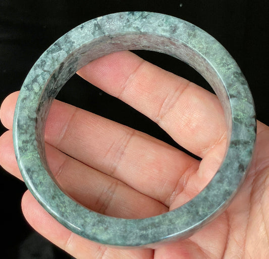Hand holding a 66.4mm certified Grade A Burmese jadeite flat style bangle in icy black and greyish with apple and moss green hues, item JB935