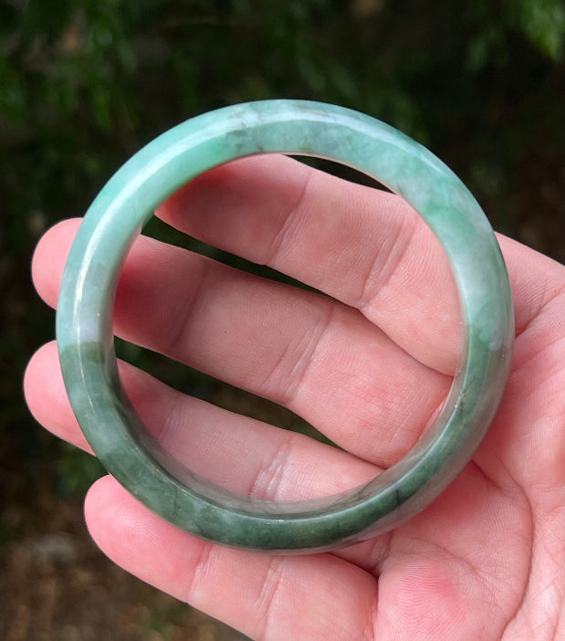 Hand holding a 58.4mm certified Grade A natural Burmese jadeite bangle in icy bluish green and moss in snow, item JB1320