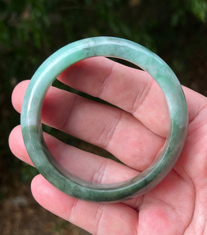 Hand holding a 58.4mm certified Grade A natural Burmese jadeite bangle in icy bluish green and moss in snow, item JB1320
