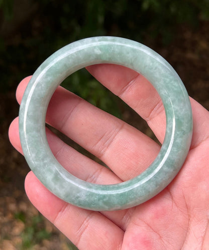 Hand holding a 54mm certified Grade A natural Burmese jadeite princess round bangle in icy celadon and apple green, item JB1318