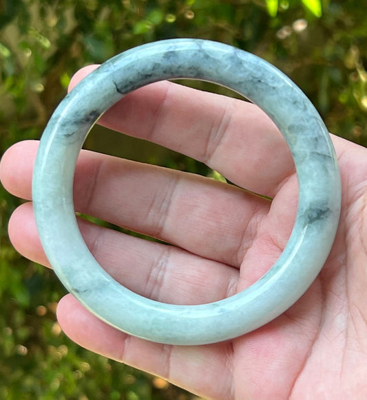 Hand holding a 64.9mm certified Grade A natural Burmese jadeite princess round bangle in icy celadon green and snow white, item JB1272