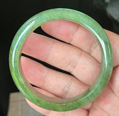 Hand holding a 58.5mm certified Grade A Burmese jadeite bangle in icy emerald and apple green hues, item JB043