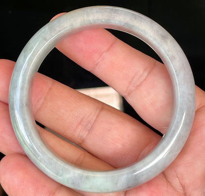Hand holding a 57mm certified Grade A natural Burmese jadeite bangle in icy grey and milky white with honey hues, item JB358