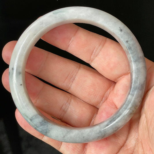 Hand holding a 66.5mm certified Grade A Burmese jadeite bangle in icy grey and milky white tones with honey hues, item JB879