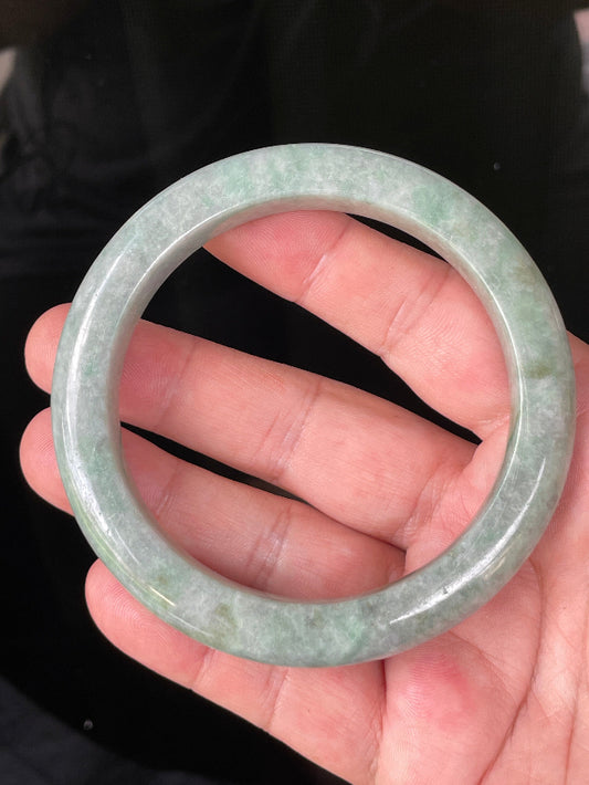 Hand holding a 66mm certified Grade A Burmese jadeite bangle in icy greyish apple and imperial green hues, item JB223