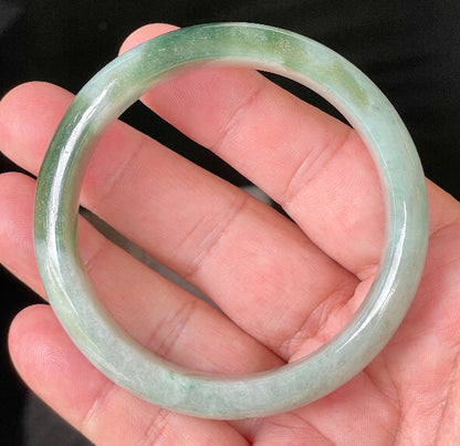 Hand holding a 58.5mm certified Grade A Burmese jadeite bangle in icy greyish milky white with apple green hues, item JB039