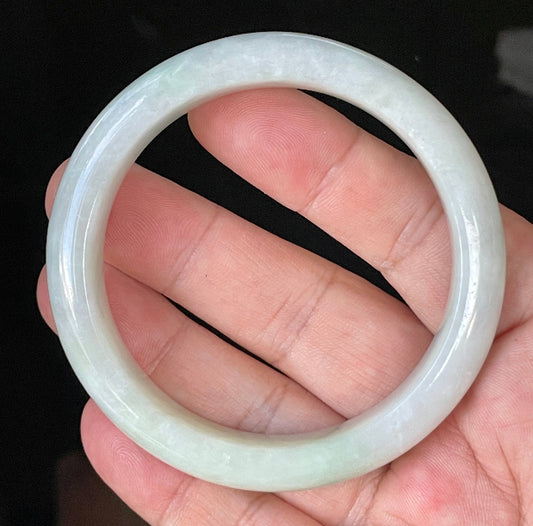 Hand holding a 58.5mm certified Grade A Burmese jadeite bangle in icy greyish milky white with apple green hues, item JB910
