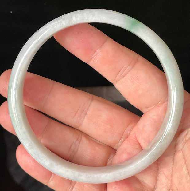 Hand holding a large 76.5mm certified Grade A Burmese jadeite bangle in icy greyish milky white with imperial green hues, item JB881