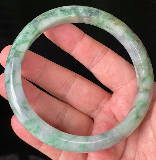 Hand holding a large 77mm certified Grade A Burmese jadeite bangle in icy imperial and apple green with greyish milky white hues, item JB933