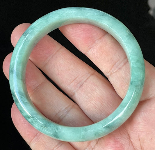 Hand holding a 56mm certified Grade A Burmese jadeite bangle in icy imperial and apple green with milky white hues, item JB914