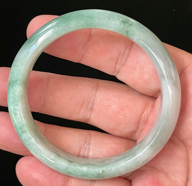 Hand holding a 58mm certified Grade A Burmese jadeite bangle in icy imperial green with greyish milky white hues, item JB004