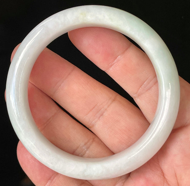 Hand holding a 57.5mm certified Grade A Burmese jadeite bangle in icy milky white with apple green hues, item JB921
