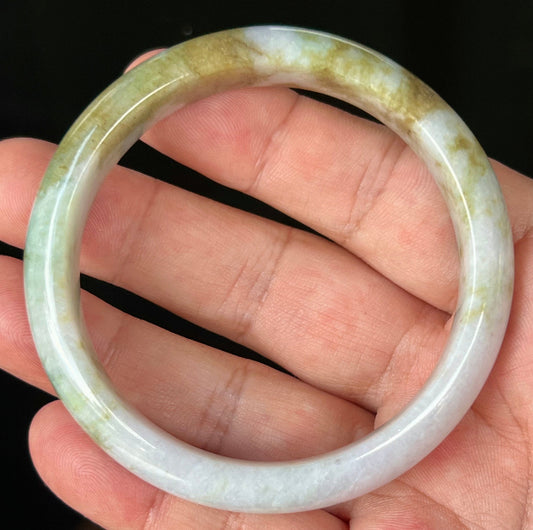 Hand holding a 58.4mm certified Grade A natural Burmese jadeite bangle in icy milky white with moss green and light brown hues, item JB535