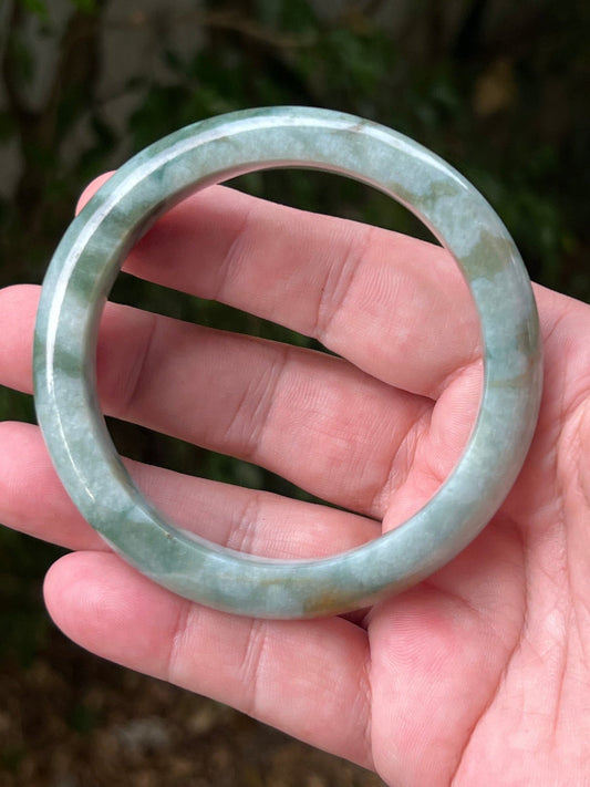 Hand holding a 62.2mm certified Grade A natural Burmese jadeite bangle in icy seafoam and moss green, item JB1313