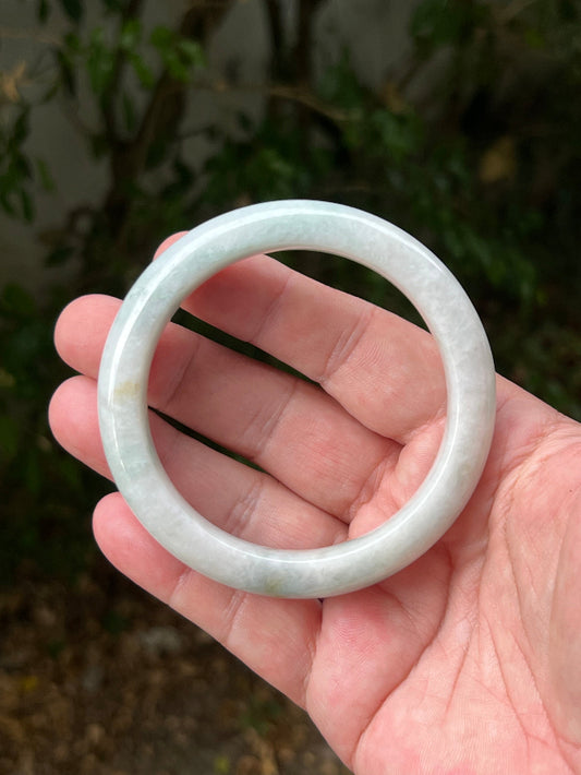 Hand holding a 62.2mm certified Grade A natural Burmese jadeite bangle in icy milky white and pale celadon green, item JB1314