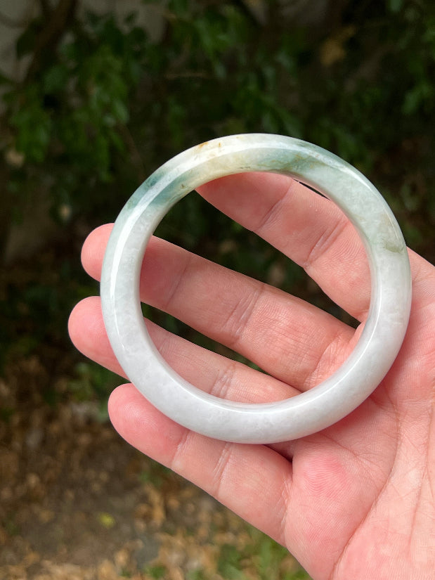 Hand holding a 62mm certified Grade A natural Burmese jadeite bangle in icy white, green, and honey, item JB1290