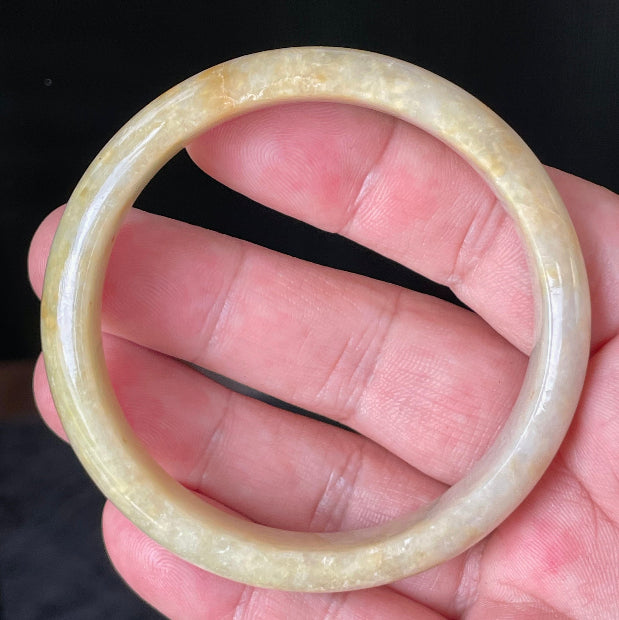 Hand holding a 58.5mm certified Grade A Burmese jadeite bangle in icy yellow and honey orange with milky white hues, item JB923