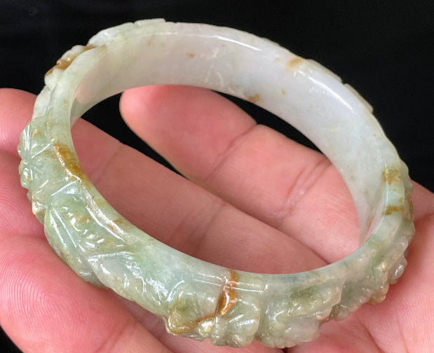 Close-up of a 57mm certified natural Grade A Burmese jadeite bangle featuring icy apple green, white, and honey hues with hand-carved floral motifs, item JB210