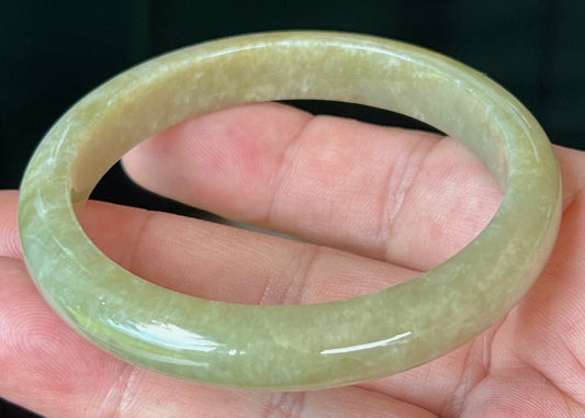 Hand holding a 56.8mm natural Grade A Burmese jadeite bangle in icy apple and forest green with light honey hues, item JB522