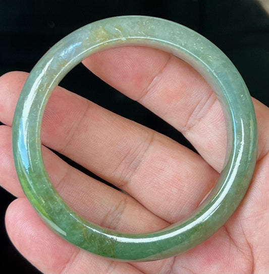 Hand holding a 56mm natural Grade A Burmese jadeite bangle in icy apple and forest green with honey and greyish hues, item JB549