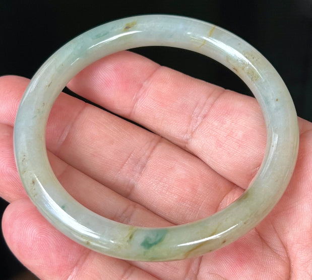 Hand holding a 58.6mm natural Grade A Burmese jadeite bangle in icy light apple green with honey and imperial green hues, item JB515