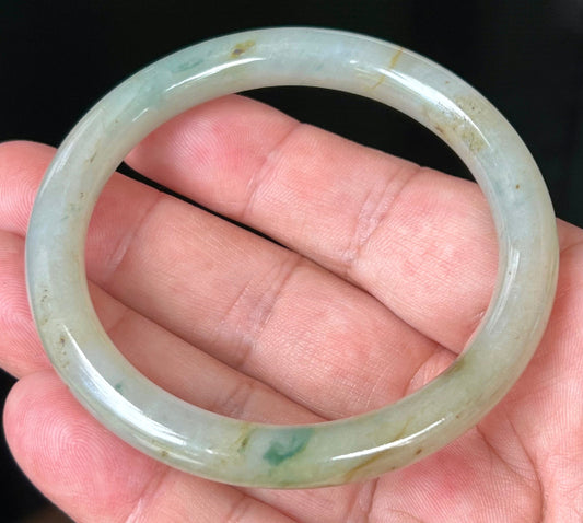 Hand holding a 58.6mm natural Grade A Burmese jadeite bangle in icy light apple green with honey and imperial green hues, item JB515