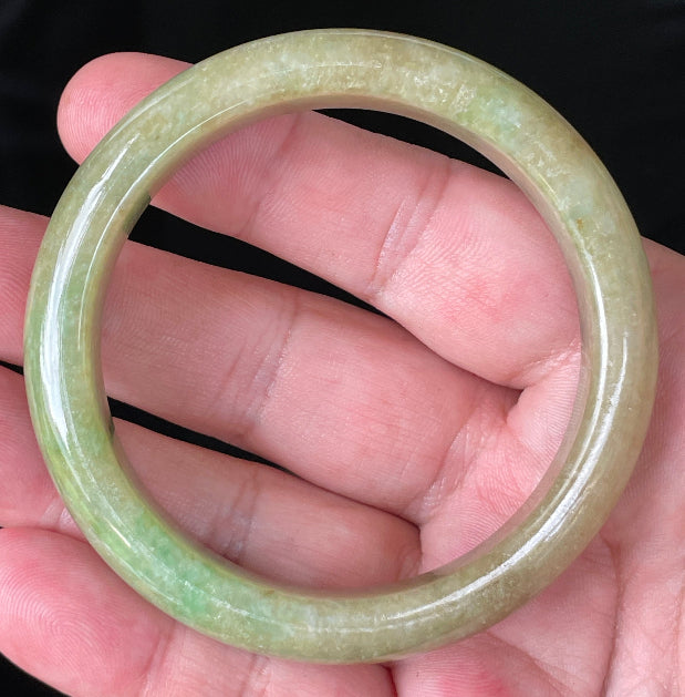 Hand holding a 56.9mm natural Grade A Burmese jadeite bangle in icy apple and imperial green with honey brown hues, item JB218